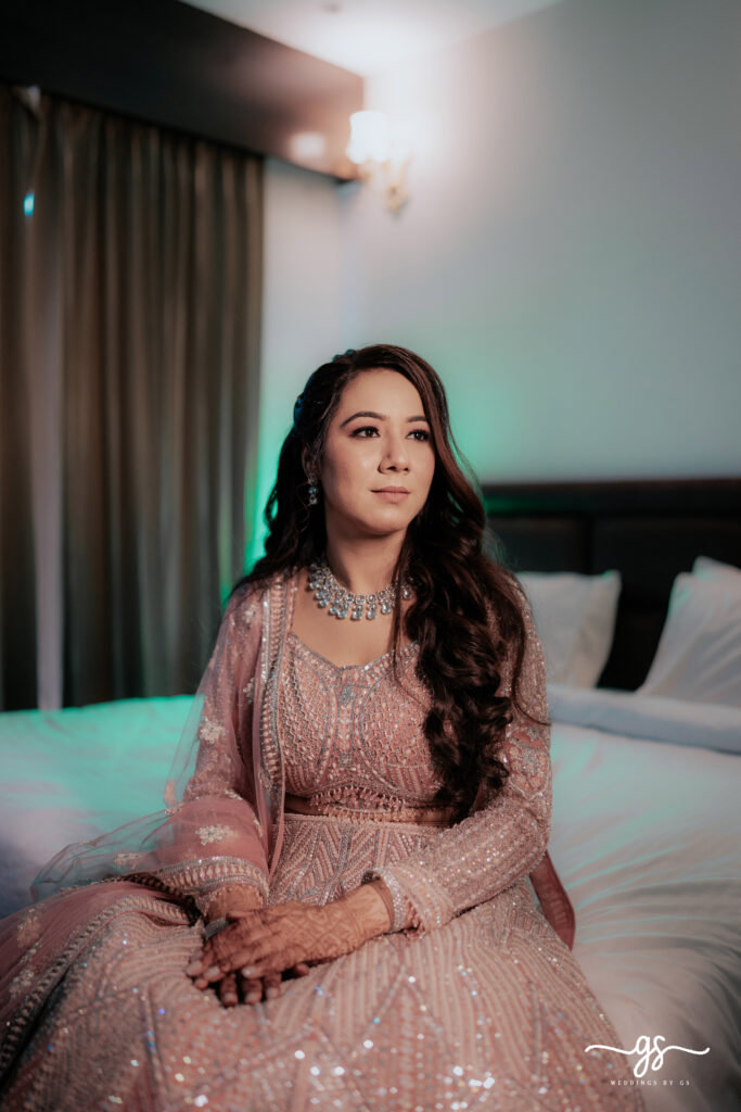 "Engagement bride sitting gracefully on bed in pastel embellished lehenga with diamond necklace – captured by Weddings by GS"
